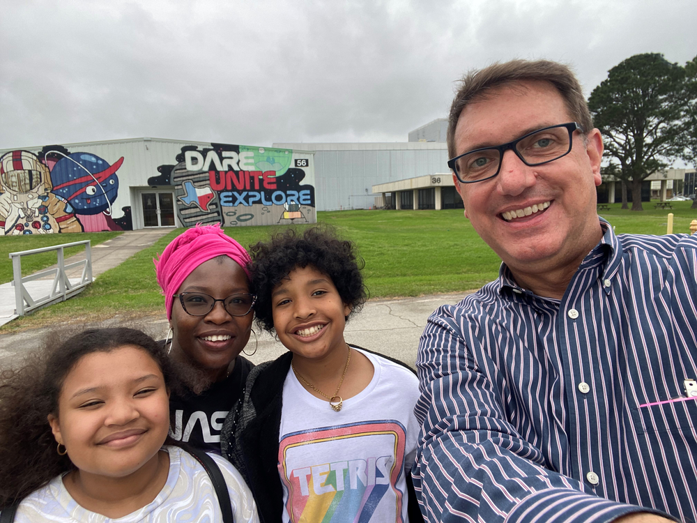 Two adults and two children take a selfie in front of a white and gray office building with a colorful mural painted on the side.